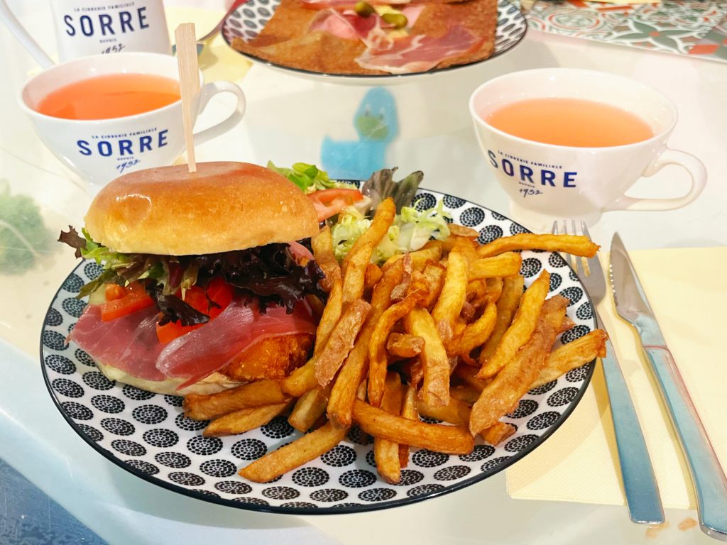 Burger L’Auvergnat avec un filets de poulet pané croustillant, Jambon cru, Forme d'Ambert, Sauce tartare, Salade, Tomate., servi avec frites, salade et tasses de cidre en arrière-plan.
